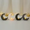 Four decorative moon-shaped candle holders with LED lights and hanging stars are displayed in a row on a surface, with blurred curtains and fairy lights in the background. Produktbild-Vorschau