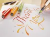 A hand is writing 'Thank you' with calligraphy pens on a white sheet of paper. Several pens are laid out on the table. Voir les images du produit