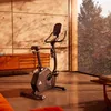 An exercise bike is placed in a modern living room with large windows overlooking a forest. Produktbild-Vorschau