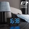 A digital alarm clock is placed on a nightstand next to a lamp and a bed. Náhled obrázku produktu
