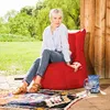 A woman is sitting on a red beanbag chair outdoors, next to a wooden structure. Produktbild-Vorschau
