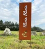 A tall, rectangular metal sign with "Moin Moin" and an anchor design, standing in a grassy field with trees and rocks in the background. Produktbild-Vorschau