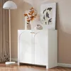 A white sideboard cabinet with arched doors and gold handles, decorated with a vase of dried leaves and a small decorative object, placed in a room with a floor lamp and curtains. Podgląd zdjęcia produktu