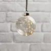 A clear glass ornament filled with small LED lights hangs against a white brick wall. Produktbild-Vorschau