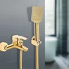 A gold-colored shower faucet and handheld showerhead are mounted on a gray tiled wall, with a blurred bathtub in the background. Podgląd zdjęcia produktu