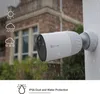 A white security camera is mounted on a white wall, with rain falling around it. In the background, there is a brick house and greenery. Icons representing sun, rain, and snow are shown on the left side of the image. Produktbild-Vorschau