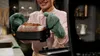 A person wearing oven mitts holds up a freshly baked loaf of bread in a dark cooking pot. Produktbild-Vorschau