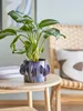 A plant in a decorative pot sits on a small wooden table next to a chair with a patterned cushion. Produktbild-Vorschau