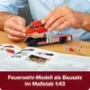 A person's hands assembling a red and silver model fire truck on a wooden table, with an open instruction book in the foreground. Produktbild-Vorschau