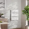A white electric towel warmer is mounted on a textured gray wall in a modern bathroom or kitchen setting. A large green plant is visible to the right, and a table with a fruit bowl is in the foreground. Anteprima dell’immagine prodotto