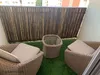 A balcony with artificial grass, two wicker chairs with cushions, and a small wicker table, all set against a bamboo fence. Náhled obrázku produktu