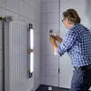 A man is working on a radiator in a tiled bathroom, with two LED work lights attached to the radiator. Produktbild-Vorschau