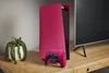 A pink PlayStation 5 console and controller are displayed on a wooden surface next to a potted plant and a television. Produktbild-Vorschau