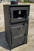 A black wood-burning stove with a glass door and a thermometer is shown outdoors on a paved surface. Produktbild-Vorschau