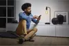 A man is sitting on the floor using his smartphone, with a smart lamp and decorative vases on a cabinet behind him. Produktbild-Vorschau