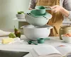 A person is holding a set of mixing bowls over a larger bowl, with ingredients like butter and pears on the counter. Produktbild-Vorschau