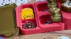 A close-up view of a pink and gold crafting kit, showing compartments with yellow circles featuring glasses, safety pins, and small star-shaped confetti. Anteprima dell’immagine prodotto