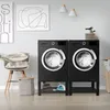 A laundry room scene with two black washing machine pedestals, each holding a washing machine. There is a chair with pillows, a laundry basket, and an iron on the floor. Náhľad obrázku produktu