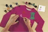 Hands painting a design onto a pink long-sleeved shirt with fabric paints. Produktbild-Vorschau