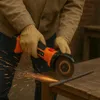 A person wearing gloves and a jacket is using a cordless angle grinder to cut a metal rod on a wooden table. Podgląd zdjęcia produktu