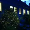 A bush decorated with warm white LED lights at night, in front of a dark house with illuminated windows. Produktbild-Vorschau