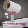 Close-up of a white track light spotlight head, showing its adjustable joint and the track it attaches to. The background is blurred, showing a room interior with furniture and lighting. Produktbild-Vorschau