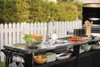 An outdoor kitchen setup with a sink, grill, and various kitchenware, set against a white picket fence and greenery. Náhled obrázku produktu