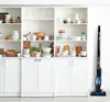 A cordless vacuum cleaner stands next to a white shelving unit filled with kitchenware. Náhľad obrázku produktu