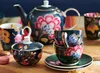A collection of decorative tea ware, including a teapot, sugar bowl, and cups, arranged on a wooden surface with macarons. Produktbild-Vorschau