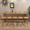 A long, live-edge wooden bar table with five stools in front of it, set against a gray wall with decorative items. Produktbild-Vorschau