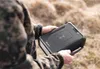 A person in camouflage clothing holds a black power bank outdoors. Produktbild-Vorschau