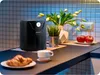 A black air fryer is placed on a kitchen counter next to a croissant and a cup of coffee. In the background, there is a vase with tulips and a potted plant. Anteprima dell’immagine prodotto