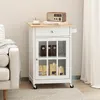 A white kitchen cart with a wooden top is shown in a room with a sofa and curtains. Podgląd zdjęcia produktu