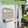 A mailbox is mounted on a white textured wall next to a white door with a garden visible in the background. Produktbild-Vorschau
