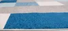 A close-up, low-angle shot of a rug with a geometric pattern of blue, gray, and white rectangles. Náhled obrázku produktu