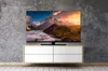 A television set displaying a landscape image is placed on a white cabinet against a textured gray wall. Produktbild-Vorschau