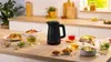 A black Bosch electric kettle is displayed on a kitchen counter with several bowls of food and cups of tea. Podgląd zdjęcia produktu
