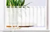 A sheer white curtain hangs in a window, partially obscuring a potted plant and a wooden surface. A few white flowers are placed on the windowsill in the foreground. Produktbild-Vorschau