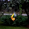 A decorative metal wind chime with a flame-like design and a glowing orb hangs from a tree branch in a garden setting. Produktbild-Vorschau