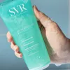 A hand holding a turquoise tube of SVR Physiopure cleansing gel with some foam. Voir les images du produit