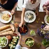 A group of people are enjoying a meal together at a wooden table, with various dishes and drinks laid out. Produktbild-Vorschau