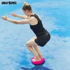 A woman is performing a squat exercise on a pink balance cushion on a blue surface. Produktbild-Vorschau