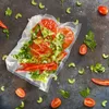 A vacuum-sealed bag containing chopped tomatoes, celery, and chili peppers, surrounded by loose ingredients on a dark textured surface. Anteprima dell’immagine prodotto