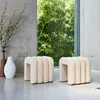 Two beige upholstered stools are placed on a concrete floor in front of a large window with lush green bamboo visible outside. A tall brown vase with dried branches and a white textured vase are to the left of the stools. Produktbild-Vorschau