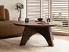 A round coffee table with a wooden finish is placed in a living room setting, next to a sofa and in front of slatted blinds. Produktbild-Vorschau
