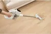 A hand holding a white cordless vacuum cleaner is shown cleaning up dirt from a wooden floor next to a couch. Podgląd zdjęcia produktu