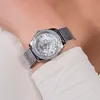 A close-up shot of a woman's wrist wearing a silver Guess watch with a mesh band and a crystal-embellished bezel. Náhled obrázku produktu
