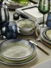 A table setting with dark blue and green glassware, plates, and bowls, along with a lit candle. Produktbild-Vorschau
