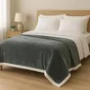 A cozy bedroom scene with a bed made up with white sheets and pillows, and a plush gray blanket draped over it. A bedside table with a lamp and a small plant is visible. Produktbild-Vorschau