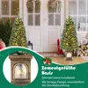 Two artificial Christmas trees with warm white LED lights are displayed in front of a house entrance with frosted glass doors and wreaths. One tree is in the foreground, partially visible, and the other is in the midground. A decorative base with lights is Podgląd zdjęcia produktu
